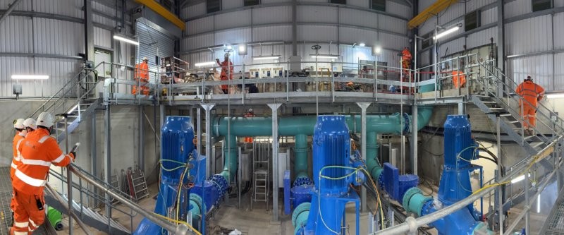 Three blue water pumps in a warehouse with working operatives at the side of the building.